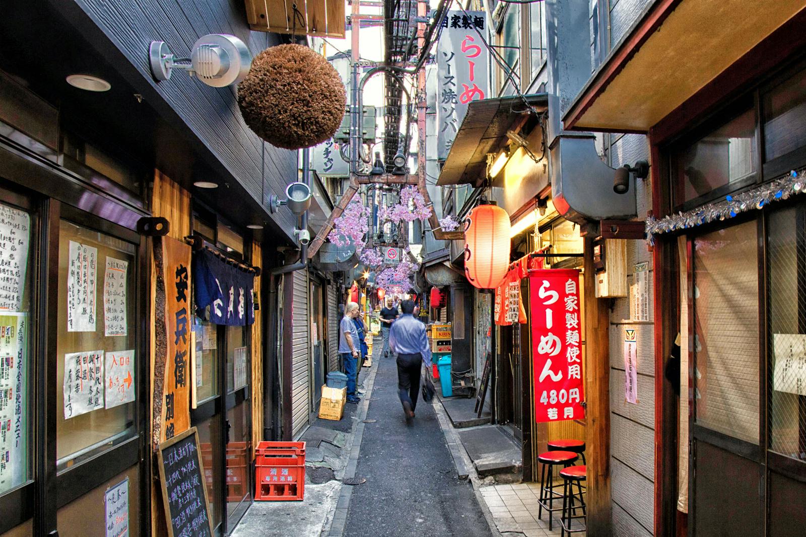 A narrow Tokyo back-street lined with small eateries and red lanterns, one sign advertising ramen for 480 yen