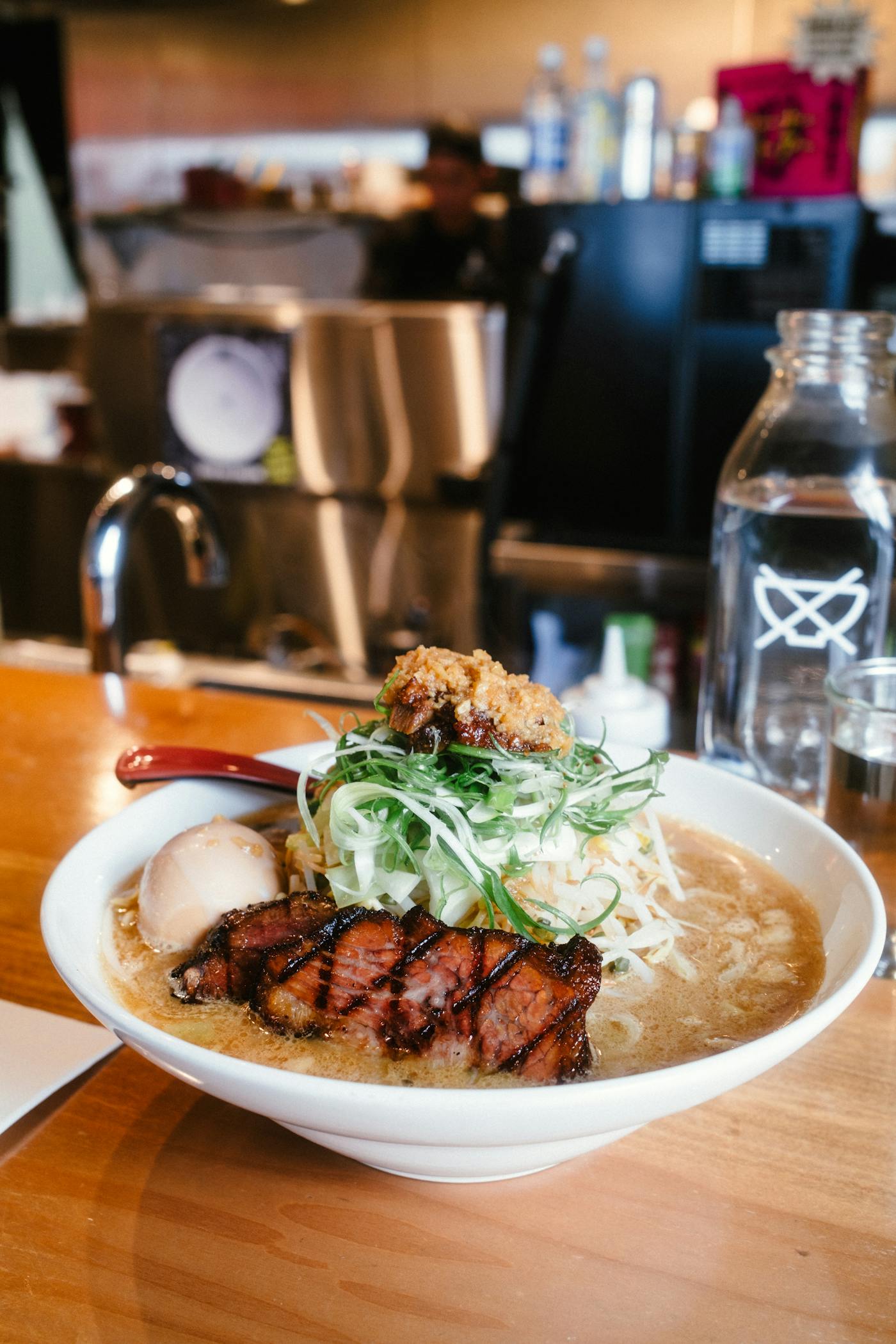 A rich bowl of Tokyo ramen at a counter shop — deep, dark broth piled with green onion and a thick slab of grilled pork