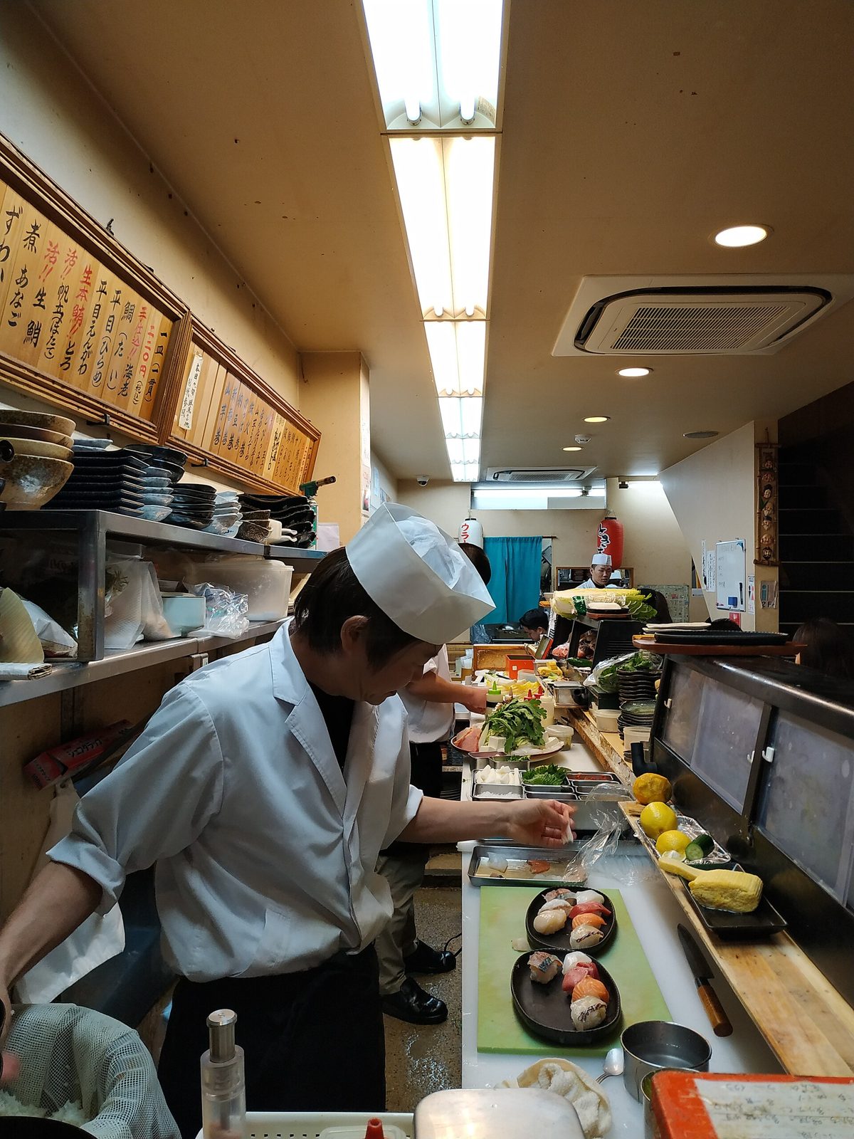 Sushi chefs making nigiri to order behind a counter with a Japanese menu board on the wall